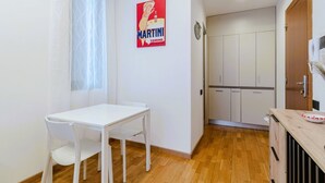 Eine kleine, helle Küche mit weißen Schränken, einer Holzausziehtafel und einem Fenster mit durchsichtigen Vorhängen.