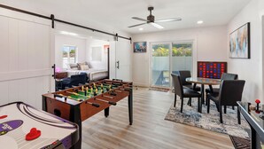 Una habitación con mesa de fútbol, comedor con sillas y mesa, ventilador de techo y puerta corrediza de vidrio que da a un balcón.