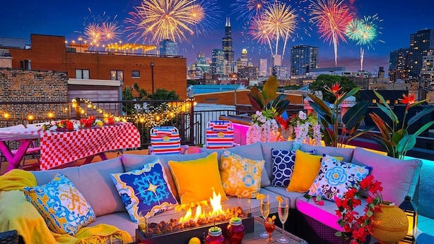 A rooftop terrace with a checkered tablecloth, a sofa with colorful cushions, a fire pit, and a city skyline with fireworks in the background. - Last-Minute Chicago home with Rooftop Views +Private HOT TUB -Garage-Sleeps 12 (Chicago)