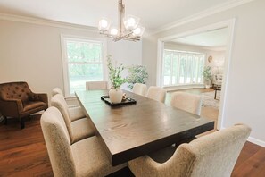 A dining area with a long table, cushioned chairs, a vase with plants, and a chandelier.