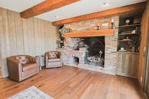 A room with a brick fireplace, two leather armchairs, a small table, and a wooden floor.