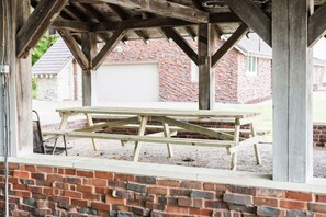 Une table de pique-nique en bois, placée sous une zone abritée, avec des murs en briques et un bâtiment blanc en arrière-plan.
