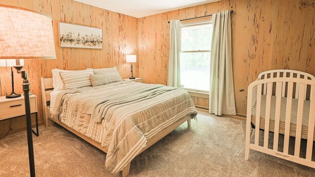 Quarto com parede de madeira, cama com manta listrada, mesa de cabeceira com abajur, berço e janela com cortinas.