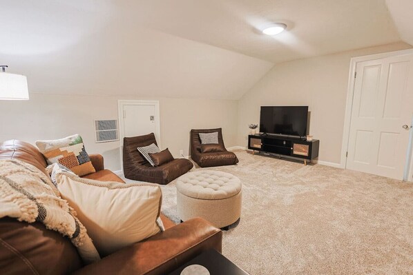 A living room with a brown leather sofa, two brown leather chairs, a white ottoman, a flat-screen TV on a black stand, and a white door in the background.