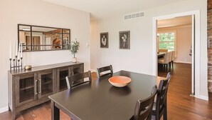Uma sala de jantar com mesa de madeira escura, cadeiras e um espelho grande acima. Há quadros nas paredes e um vaso de planta sobre a mesa.