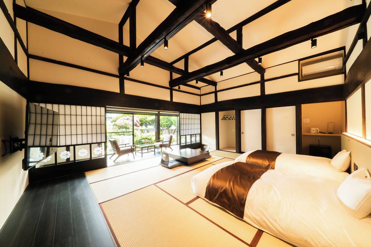 Una habitación tradicional japonesa con suelo de tatami, una cama grande, una puerta corredera que da a un balcón y vistas a la vegetación.