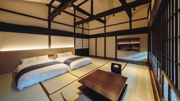 A traditional Japanese-style room with tatami flooring, a low wooden table, and a bed with two pillows. - HAPPOYA (Hachinohe)