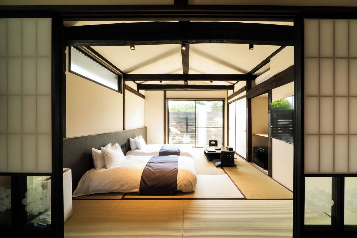Un dormitorio de estilo japonés tradicional con suelo de tatami, una cama grande con ropa de cama blanca y vistas al exterior mediante una puerta corredera.