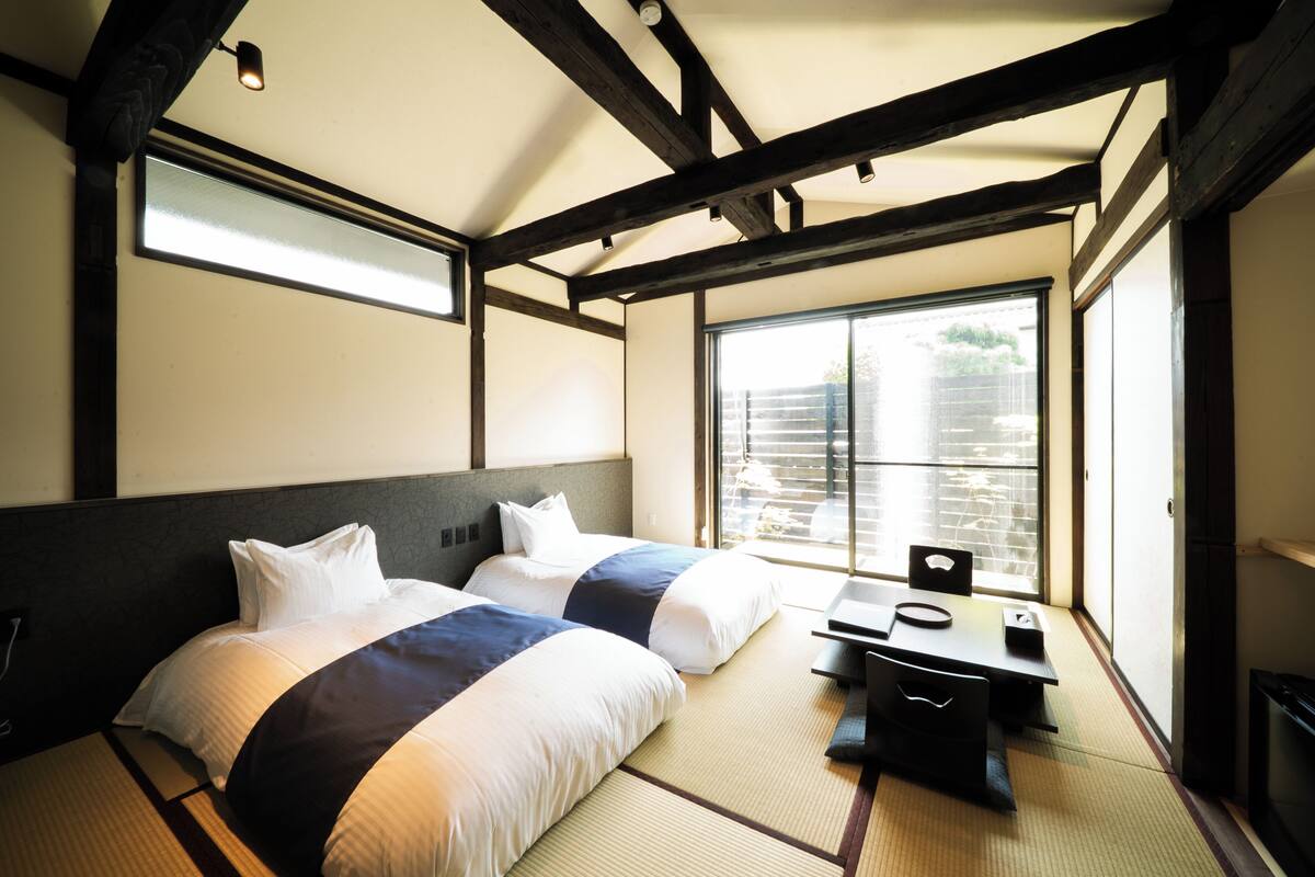 Una habitación tradicional japonesa con suelo de tatami, dos camas con ropa de cama blanca y azul, una mesita y una puerta corredera que da a un balcón.