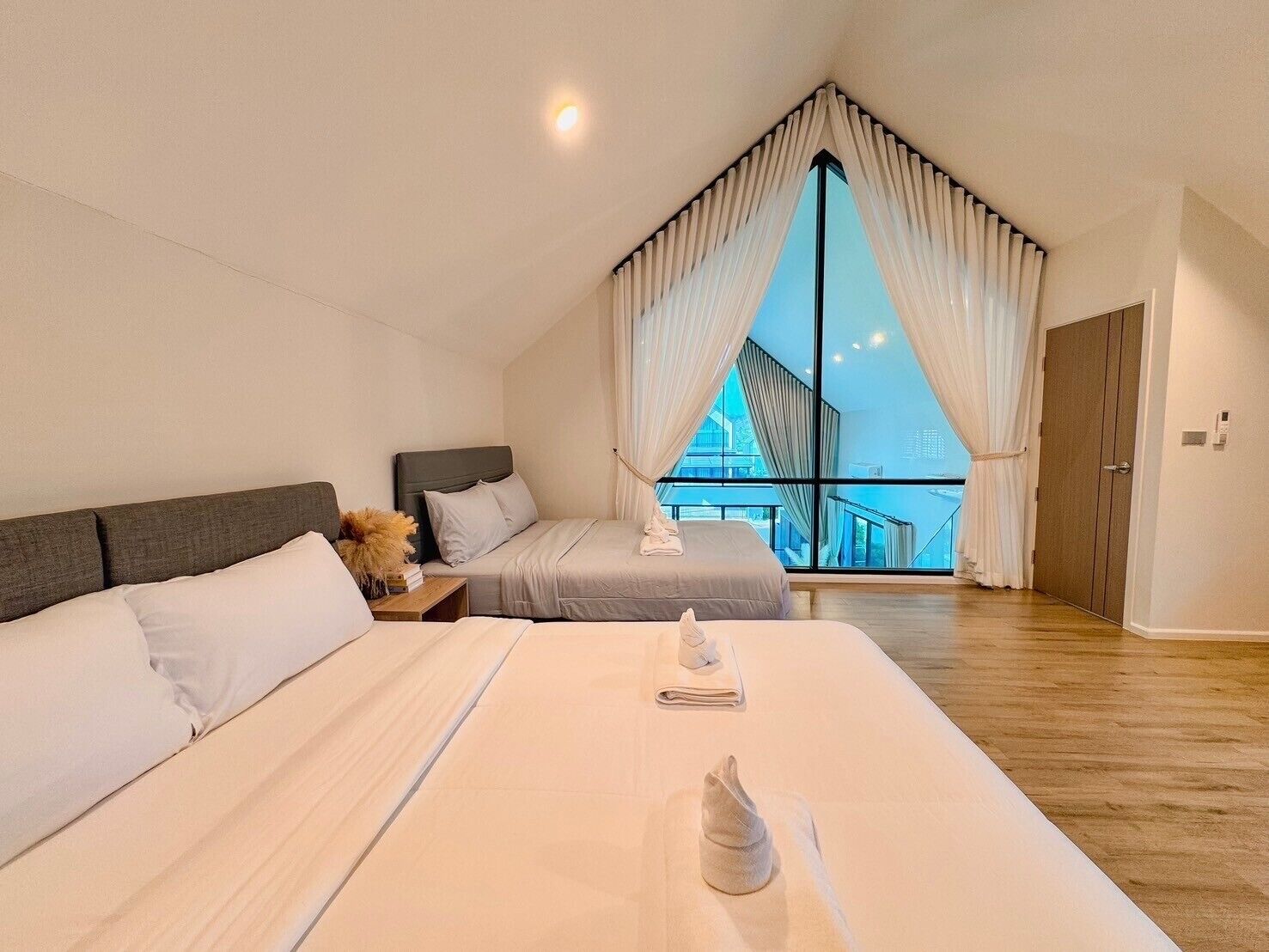 A modern bedroom with a large bed, a smaller bed, and a view of a building through sheer curtains.