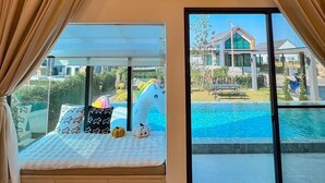 A bedroom with a large bed, a view of a swimming pool, and a house in the background.