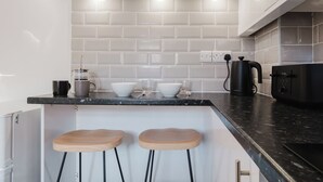 A modern kitchen with white subway tiles, a black countertop, two wooden stools, a coffee maker, and a black appliance. - Corner House - Bright 2BD in Cheltenham (Cheltenham)