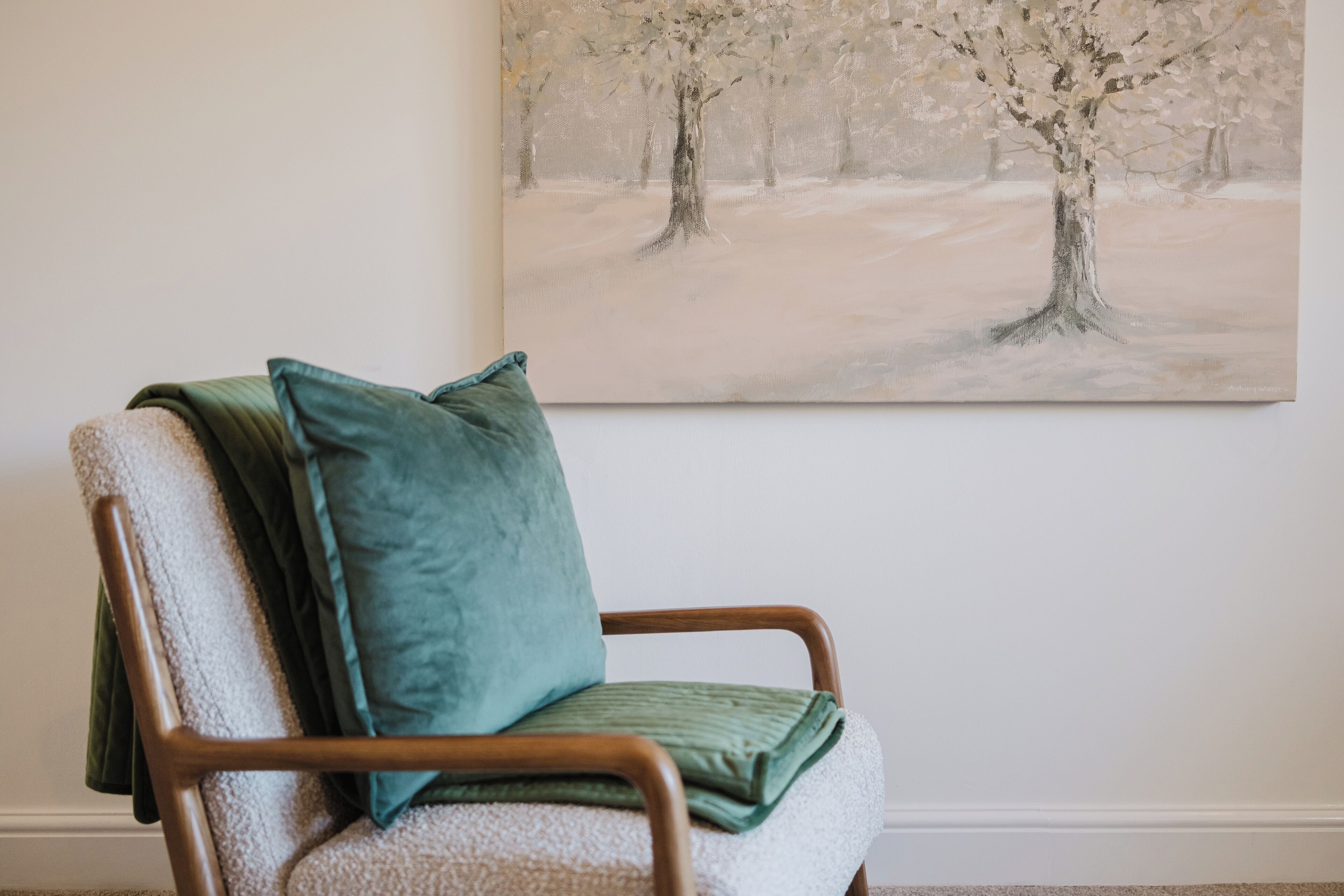 A chair with a green cushion and a blanket, positioned in front of a painting of trees on the wall.