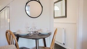 A dining area with a round table set for two, featuring white plates, glasses, and a bowl. There are two wooden chairs, a black-framed mirror on the wall, and a radiator below. - Corner House - Bright 2BD in Cheltenham (Cheltenham)