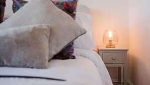 A neatly made bed with a white comforter and several pillows, including a large beige one and a colorful floral-patterned one. There is a bedside table with a lamp and a small drawer. - Corner House - Bright 2BD in Cheltenham (Cheltenham)