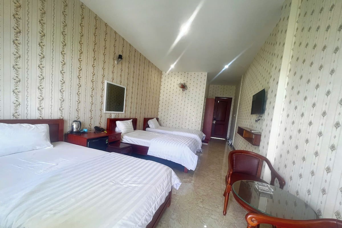 Ein Hotelzimmer mit drei Betten, einem Schreibtisch mit Telefon, einem Sessel und einem kleinen Tisch.