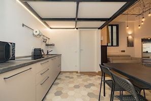 Een kleine keuken met een magnetron, broodrooster en koffiezetapparaat, een eethoek met tafel en stoelen, en een bank op de achtergrond.