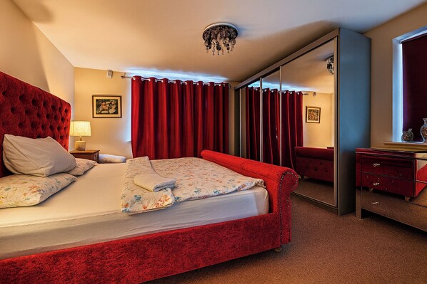 A hotel room with a large bed, red curtains, a red upholstered headboard, a bedside table with a lamp, and a mirror.