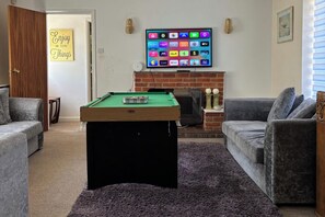 A living room with a pool table, a flat-screen TV mounted above a fireplace, and two grey sofas.