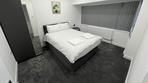 A modern bedroom with a large bed, a bedside table, a nightstand, a window with blinds, a radiator, and a potted plant on the wall.