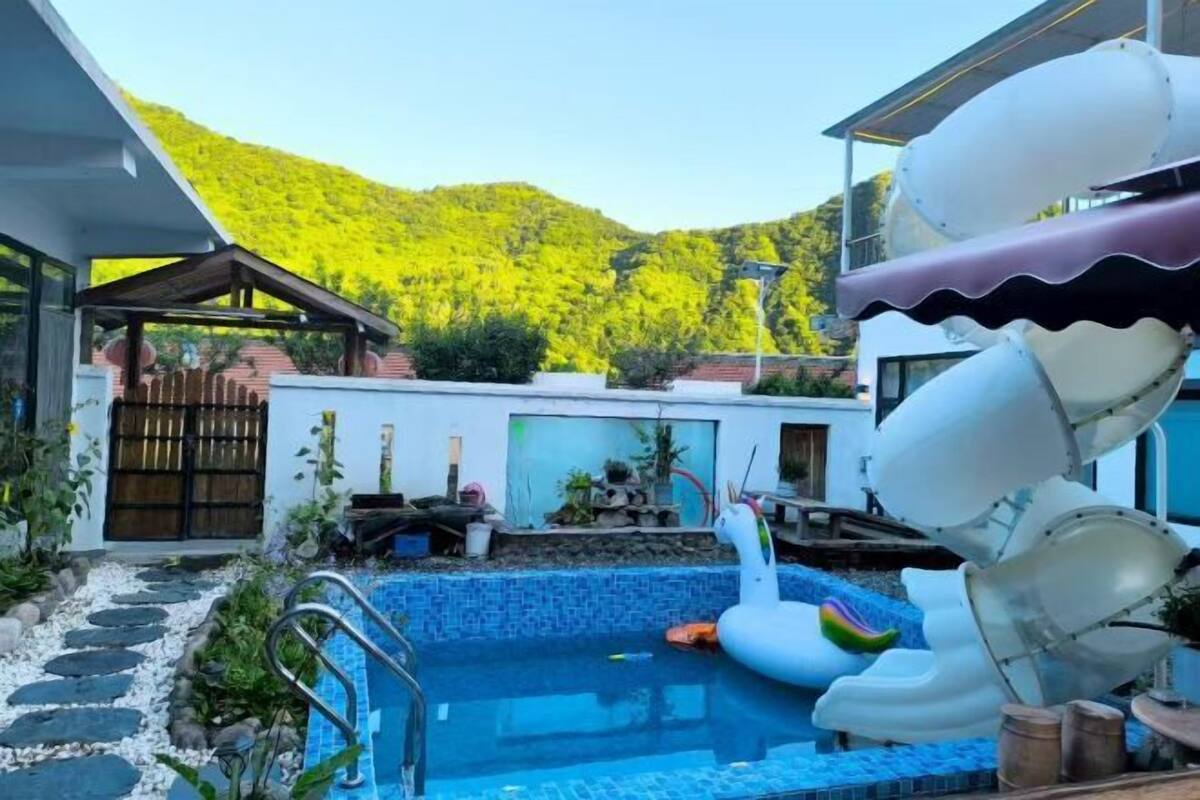 A swimming pool with a slide, surrounded by a garden and a mountain backdrop.