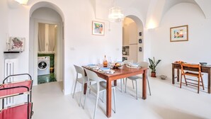 Un coin repas avec une table en bois, des chaises blanches et un lustre. Des œuvres d’art encadrées ornent les murs, et on aperçoit une cuisine au fond.