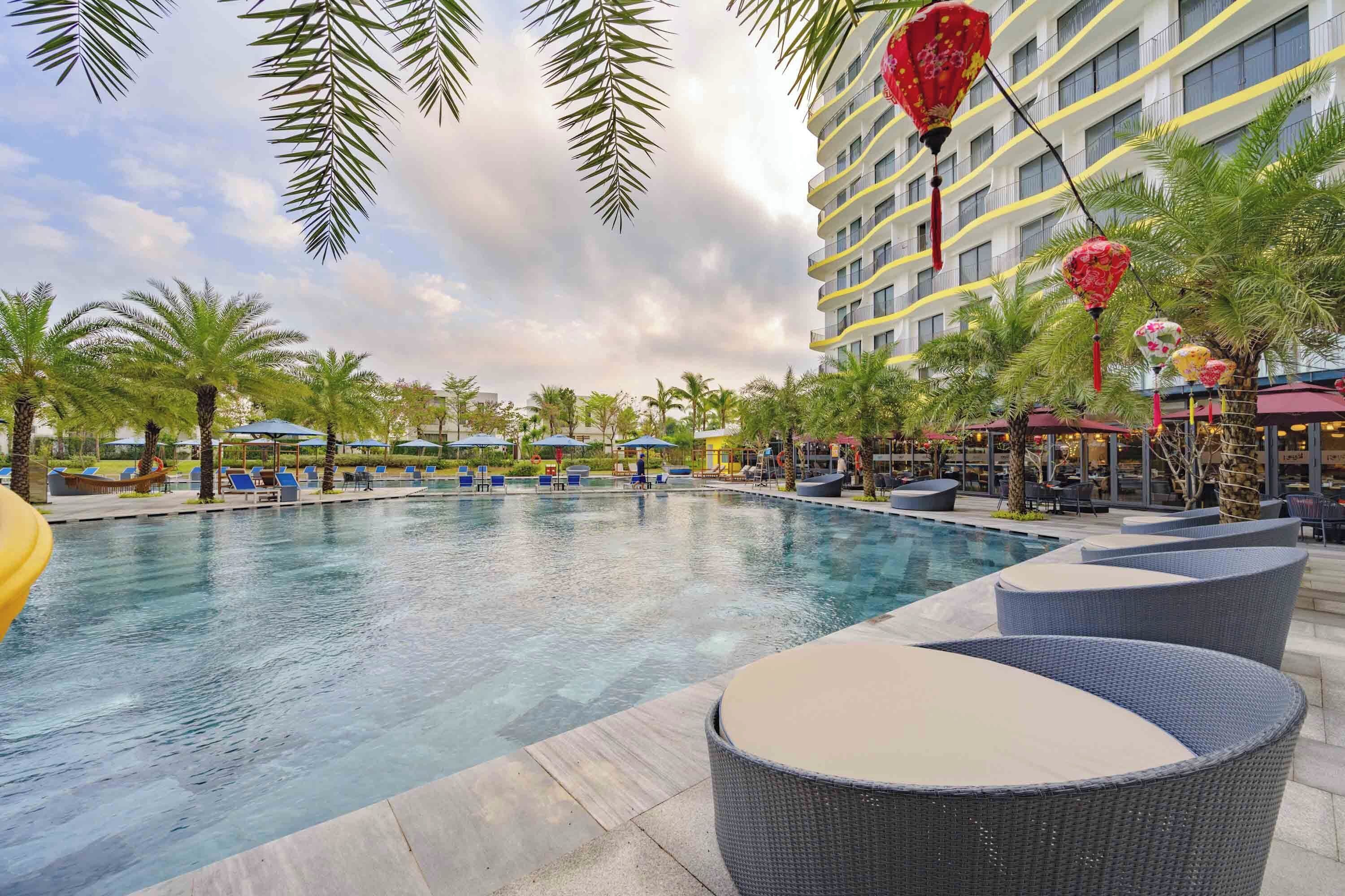 A swimming pool with lounge chairs and umbrellas, surrounded by palm trees and a hotel building.