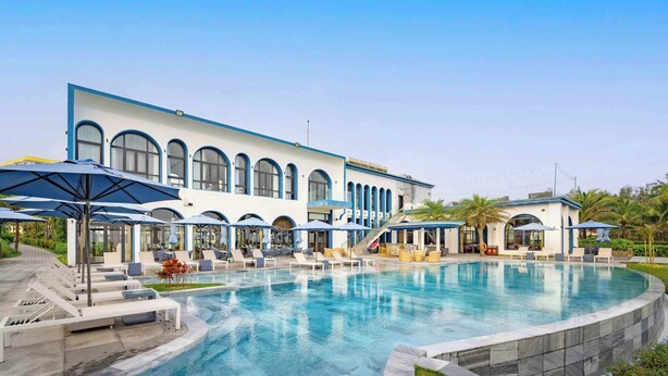 A pool area with lounge chairs and umbrellas, a building with arched windows, and a clear sky. - Aqua Vista Hoi An Beachfront Villas (Da Nang)