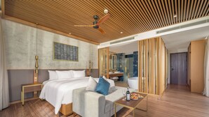 A modern hotel room with a bed, a sofa, a small table with a bottle and glasses, a ceiling fan, and a bathroom visible through a sliding door. - Aqua Vista Hoi An Beachfront Villas (Da Nang)