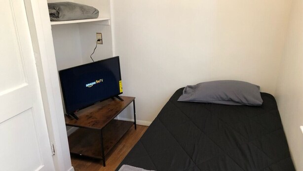 Quarto pequeno com cama, televisão sobre suporte de madeira e estante com cobertor.