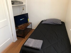 Una habitación pequeña con una cama, un televisor sobre un soporte de madera y un estante con una manta.