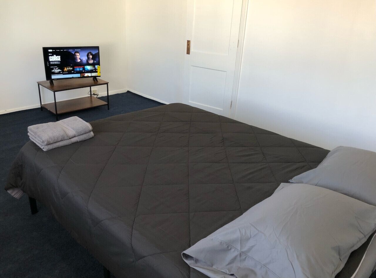A single bed with a dark quilt and two pillows, a television on a stand, and a door in the background.