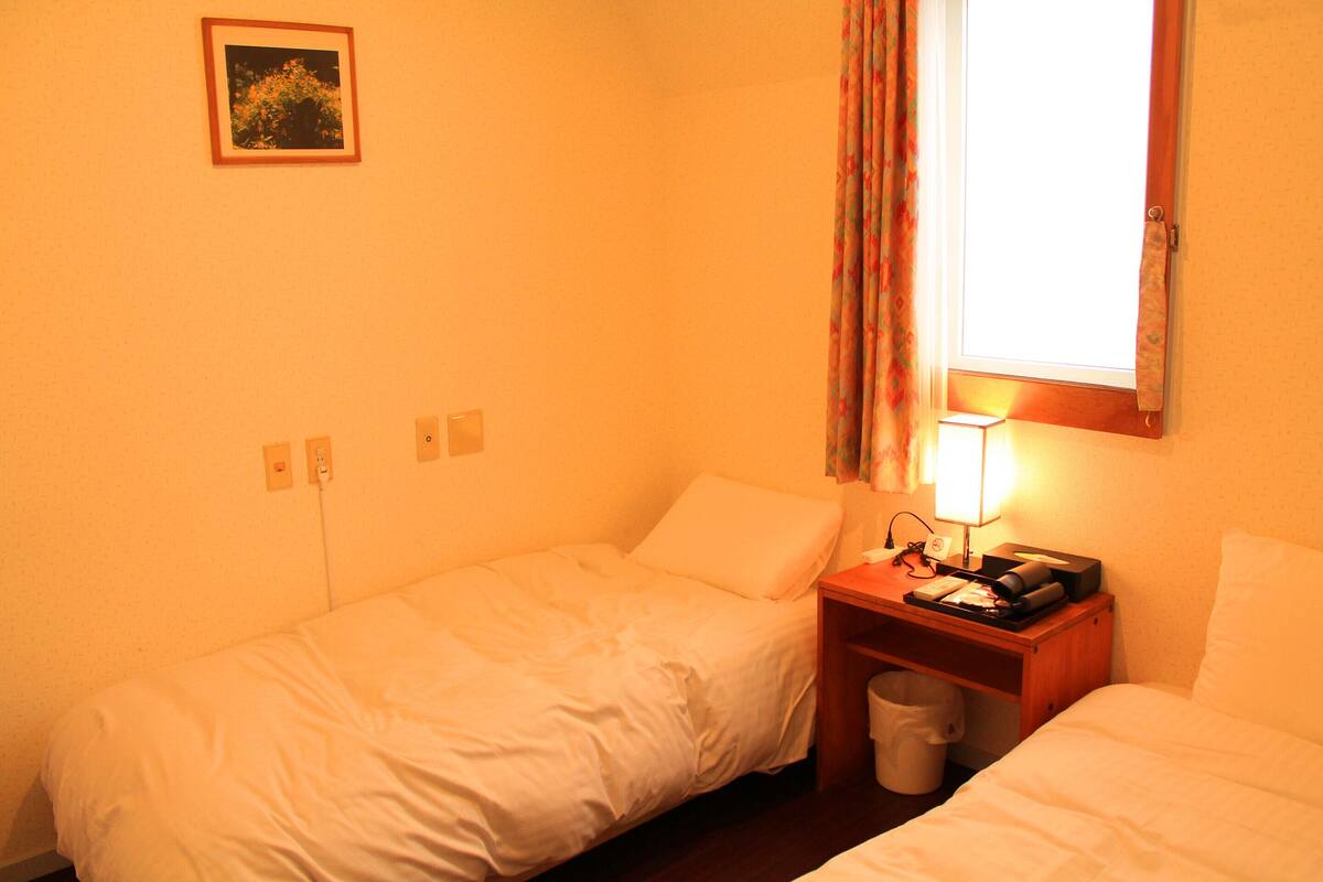 Une petite chambre d’hôtel avec deux lits simples, une table de chevet avec une lampe, une poubelle et une fenêtre avec des rideaux.