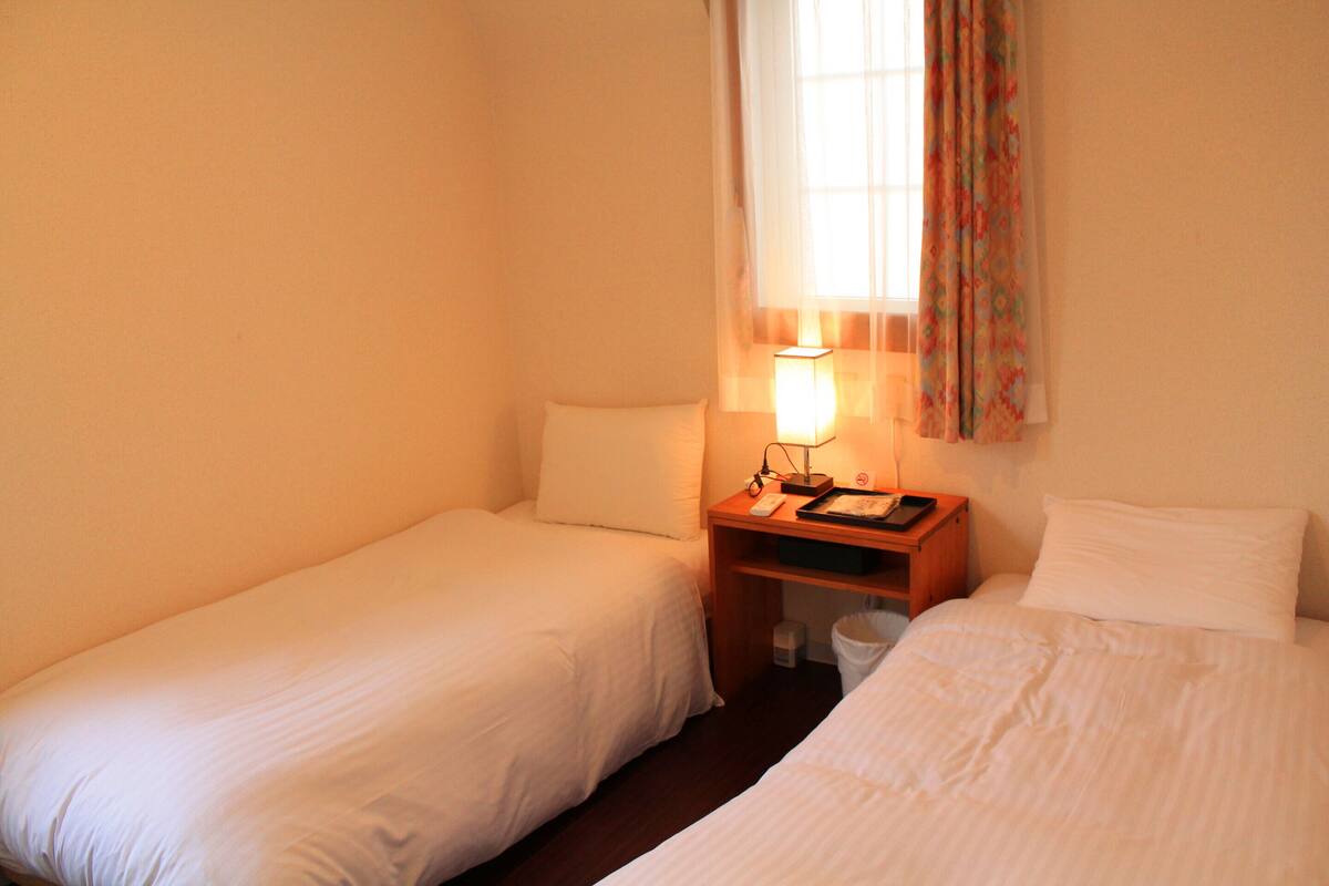 Une chambre avec deux lits simples, une table de chevet avec une lampe et une fenêtre avec des rideaux.
