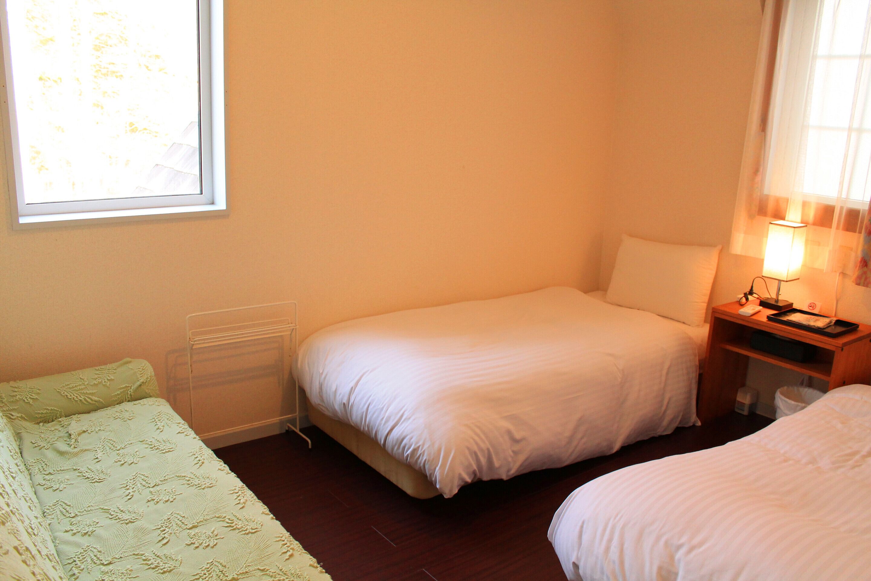 A small, single-room hotel room with a single bed, a bedside table with a lamp, and a window providing natural light.