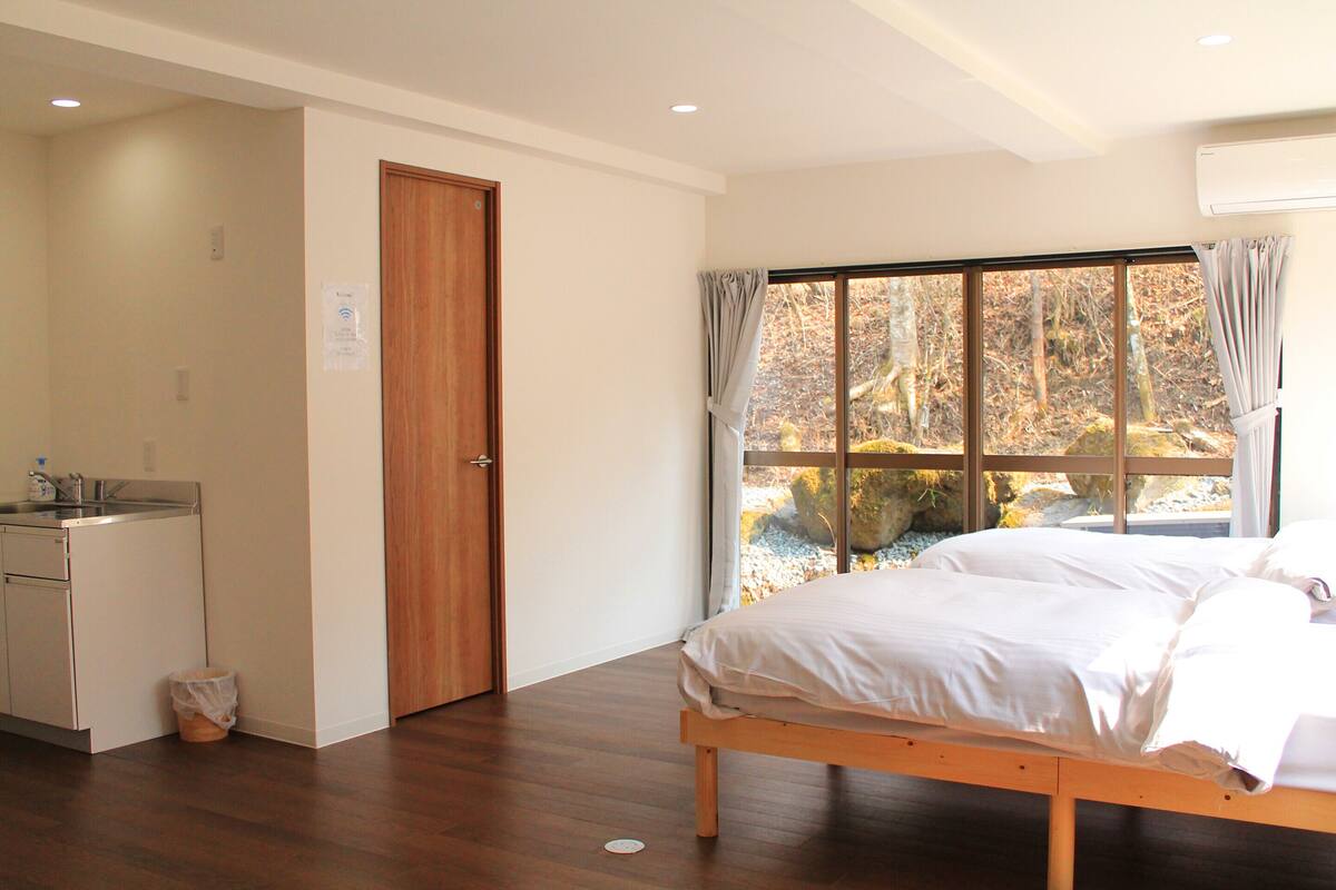 Une chambre avec un lit, une fenêtre avec des rideaux, une porte en bois et un petit coin cuisine.