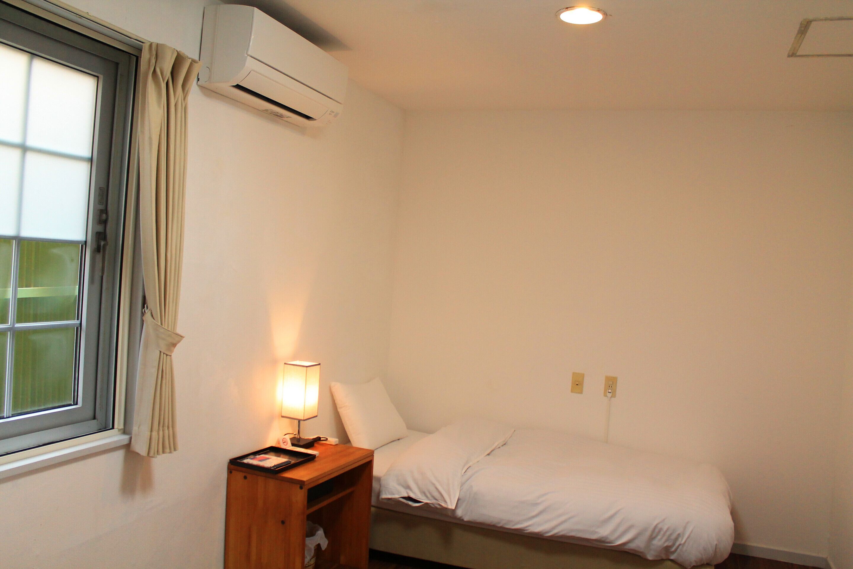 A small, clean room with a single bed, a bedside table with a lamp, and an air conditioning unit mounted on the wall.