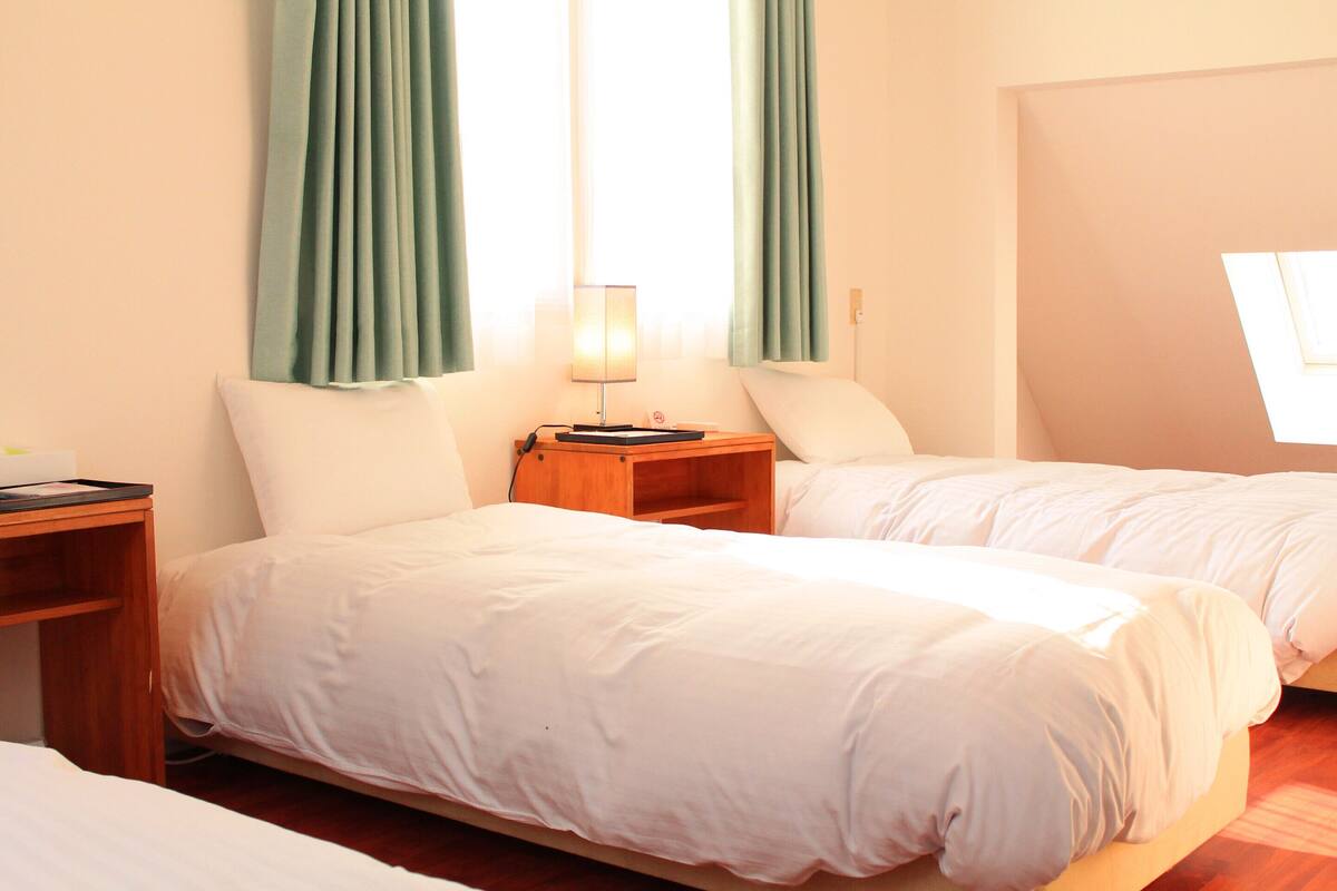Une chambre d’hôtel avec deux lits, une table de chevet avec une lampe et une fenêtre avec des rideaux.
