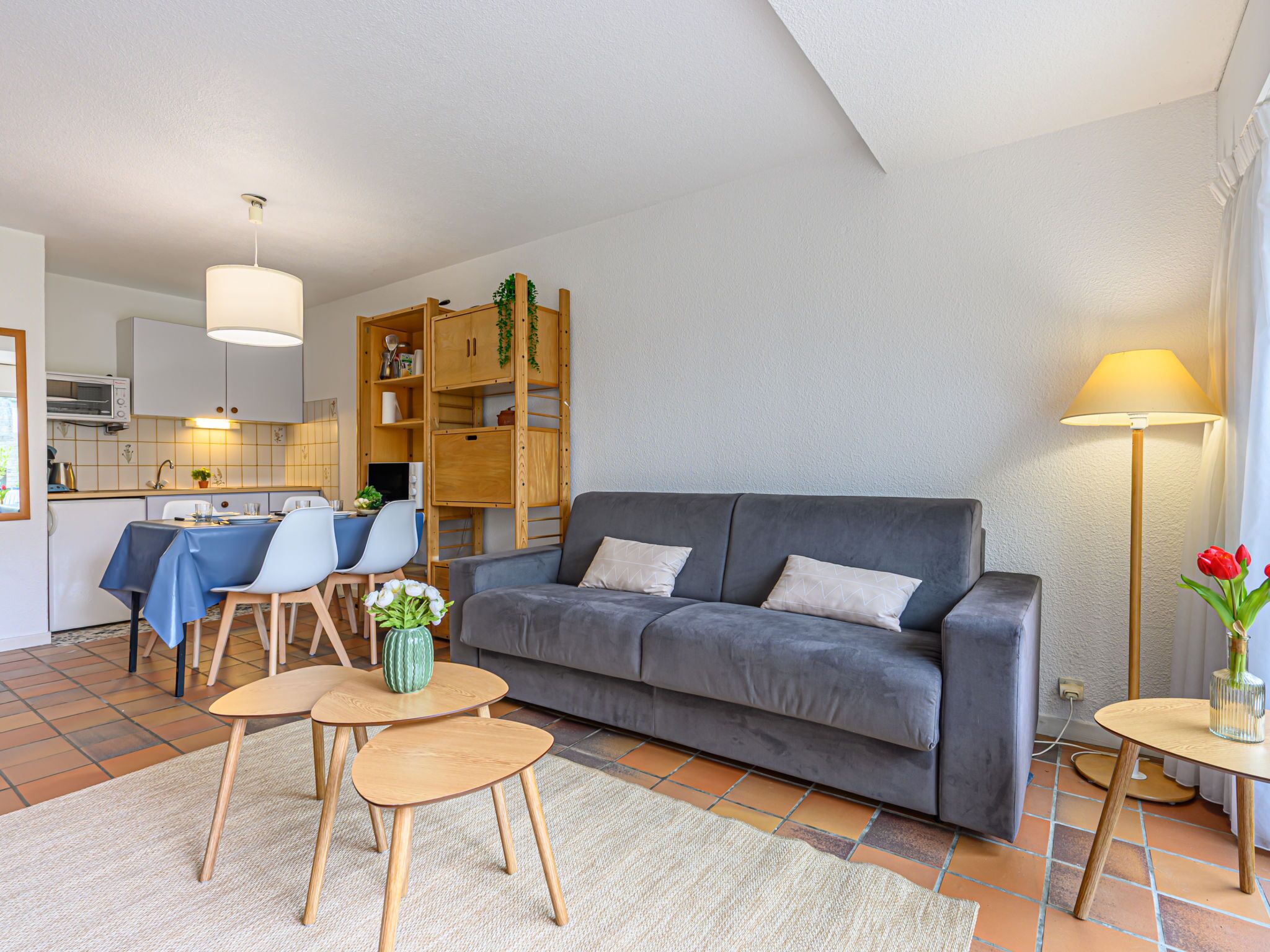 A modern living room with a grey sofa, wooden coffee table, and chairs. There is a kitchen area with a dining table and chairs, and a wooden shelving unit with plants.