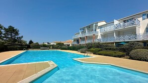 A swimming pool with a clear blue water, surrounded by a beige deck. There are white buildings with balconies in the background. - L'Orée du Golf by Interhome (Pornic)
