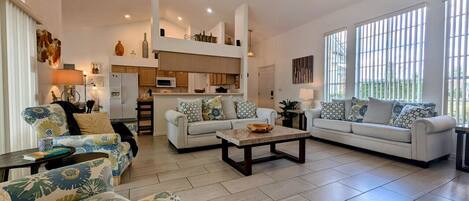 A spacious living room with a high ceiling, a ceiling fan, and a kitchen in the background.