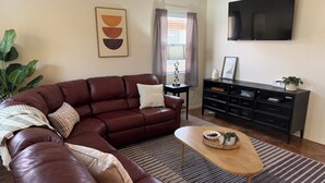 A living room with a brown leather sofa, a wooden coffee table, a black entertainment unit, and a large flat-screen TV mounted on the wall.
