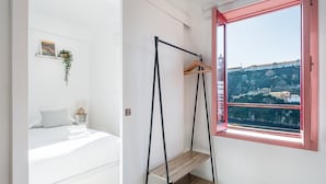 Ein Schlafzimmer mit einem Bett, einem Nachttisch, einer hängenden Pflanze, einem hölzernen Kleiderbügel und einem Fenster mit Blick auf eine Hügellandschaft.