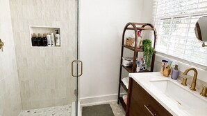Un baño con cabina de ducha de vidrio, tocador con lavabo y ventana con persianas.