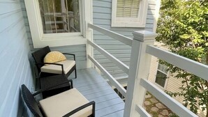 Una terraza de madera con barandilla blanca, dos sillas de mimbre con cojines y una mesita.