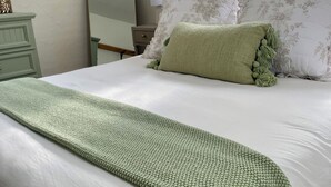 Una cama bien hecha con un plaid verde tejido, almohadas blancas con motivos florales y una mesita de noche con un espejo y una pequeña planta.