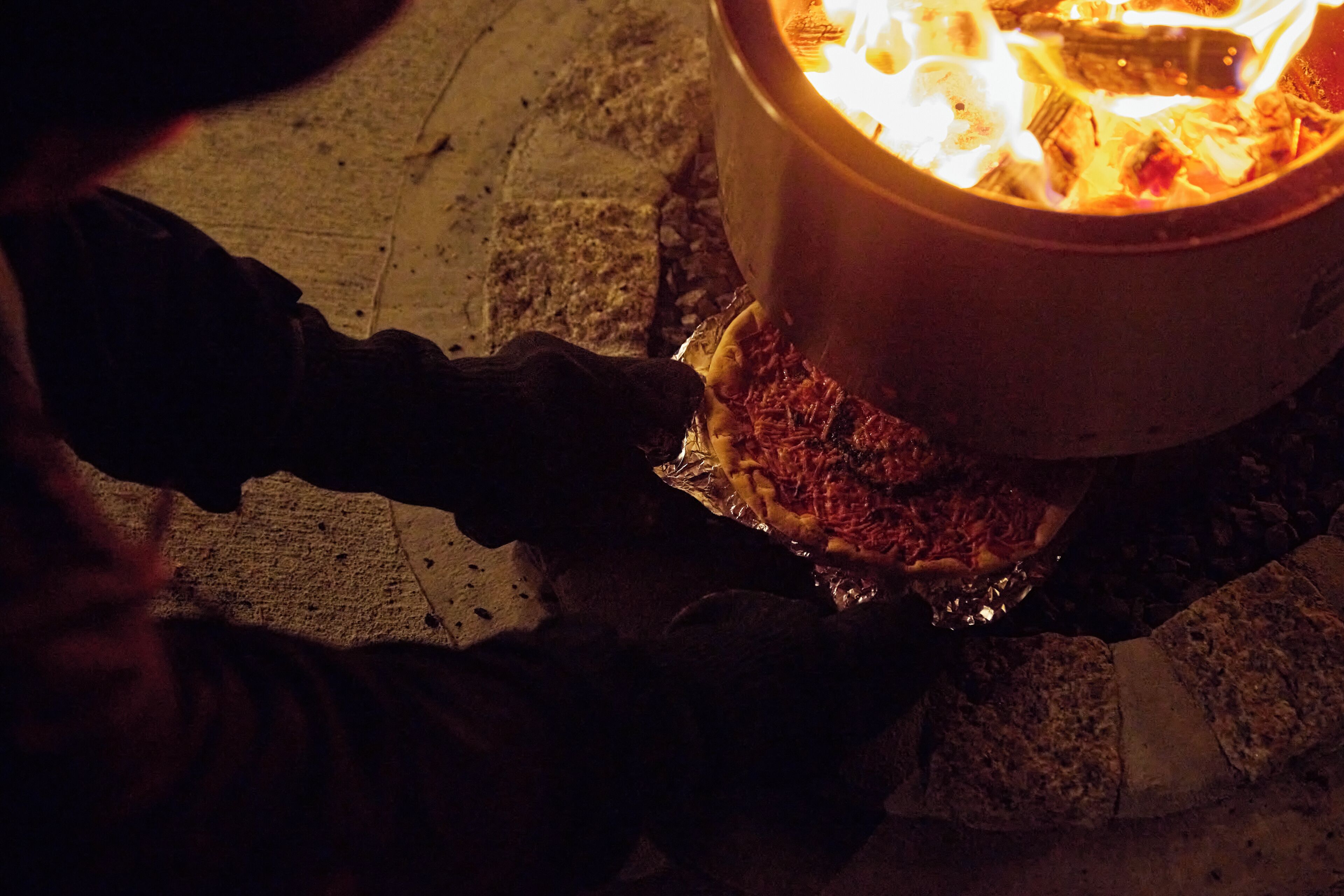 Eine Person bereitet Essen über einem offenen Feuer in einer von Steinmauern umgebenen Außenbereiches zu.
