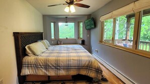 Ein Schlafzimmer mit Bett, Deckenventilator, Fernseher und Fenstern mit Blick auf Bäume.