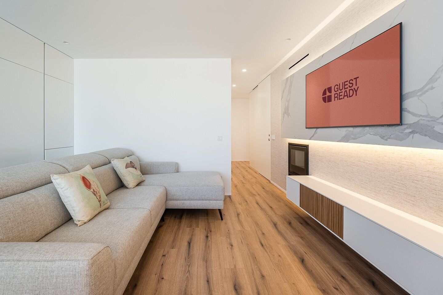Ett modernt hotellrum med en grå soffa, trägolv, en väggmonterad TV och en skylt med texten ”KLART FÖR GÄSTER”.