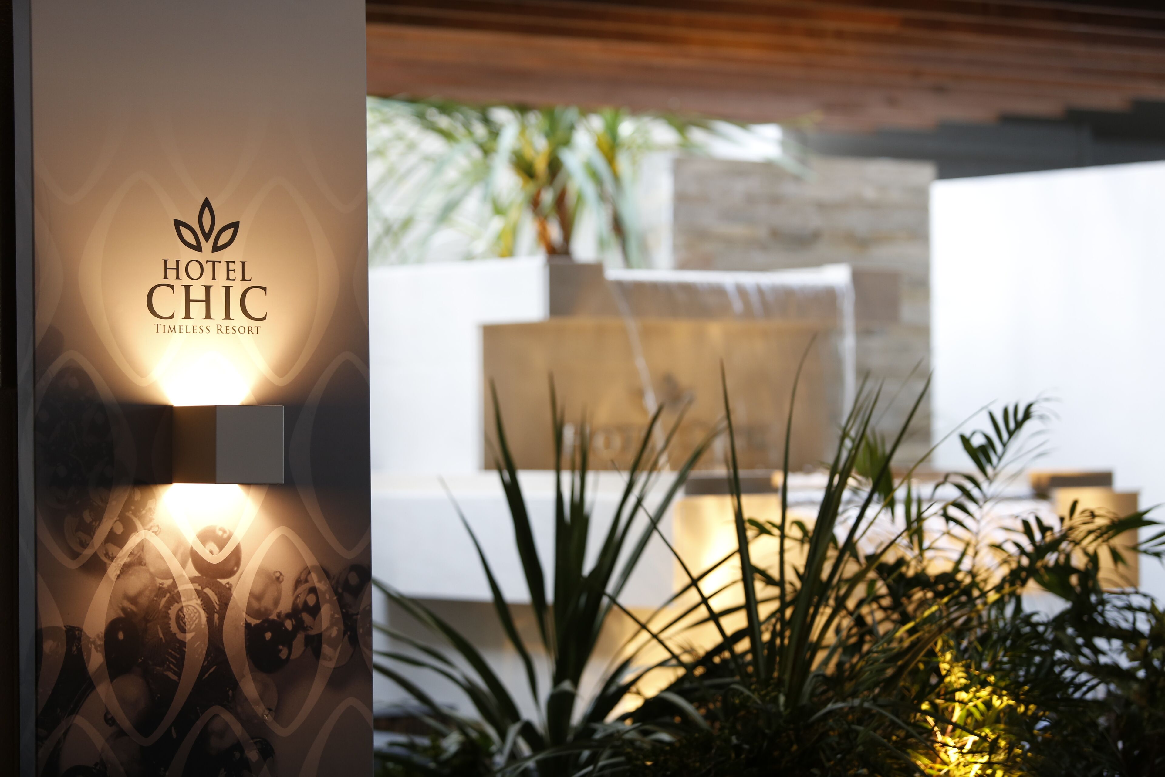 Et moderne hotelindretning med et logo på væggen, et godt oplyst siddeområde og indendørs planter.